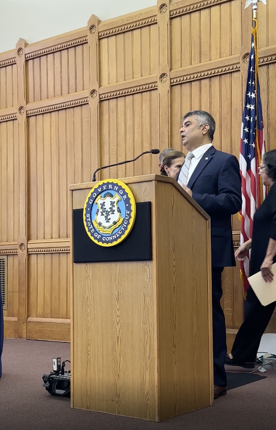 Rajeev Pahuja speaking at a podium bearing the seal of the Governor of Connecticut