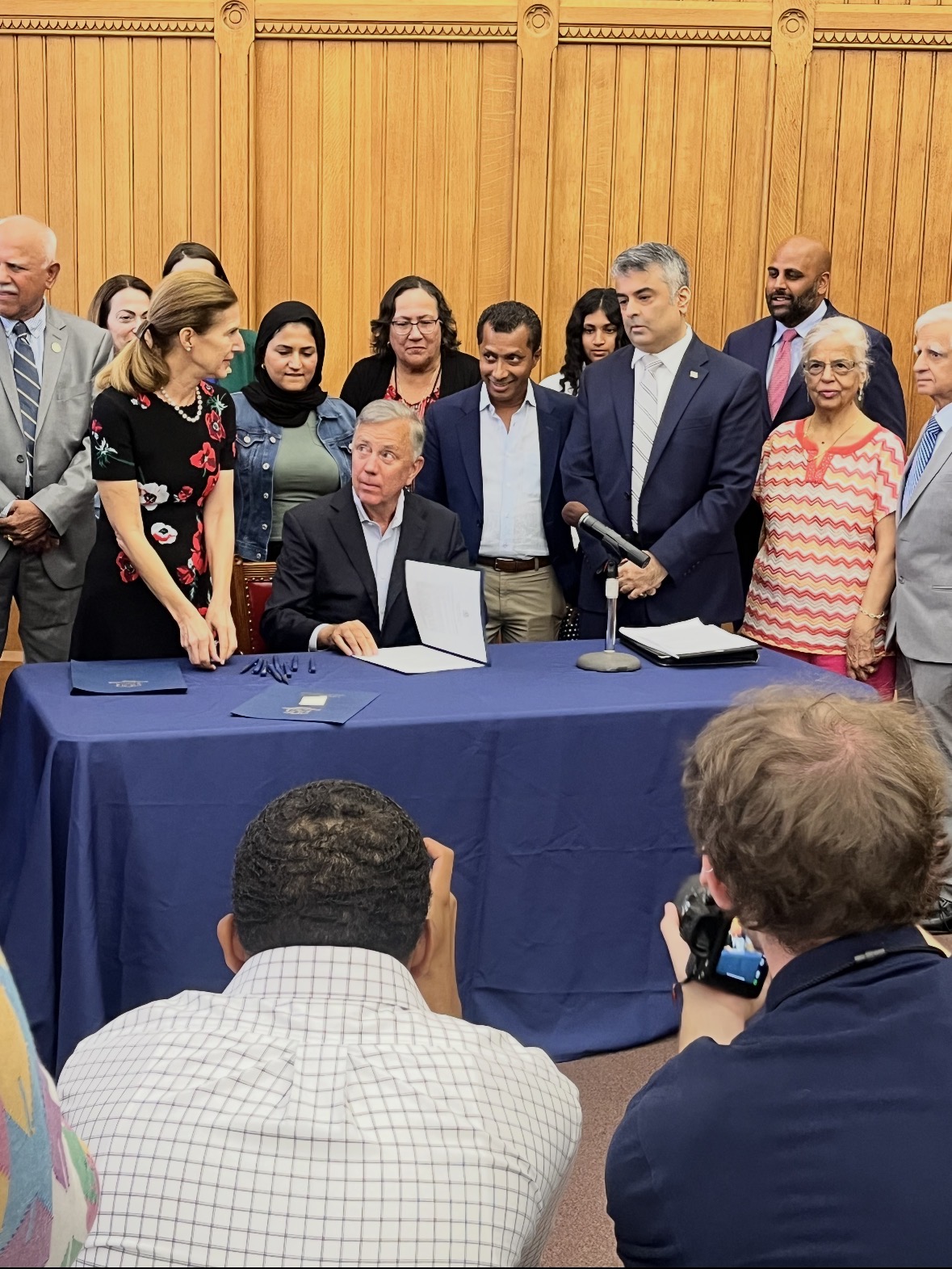Governor Ned Lamont seated signing Senate Bill 1519 with Rajeev Pahuja standing behind him among community members