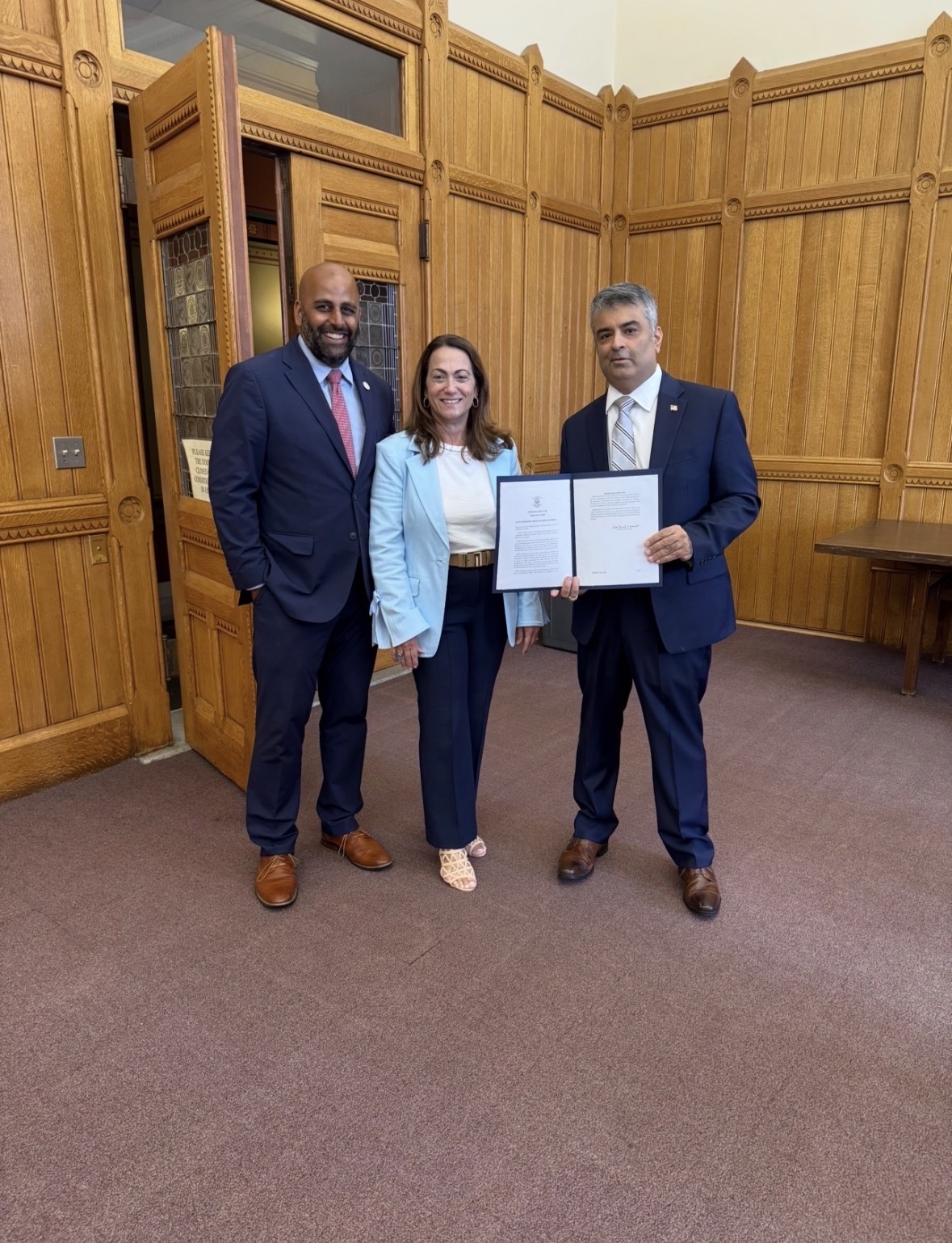 Rajeev Pahuja holding an official document with two Connecticut leaders in a wood-paneled room