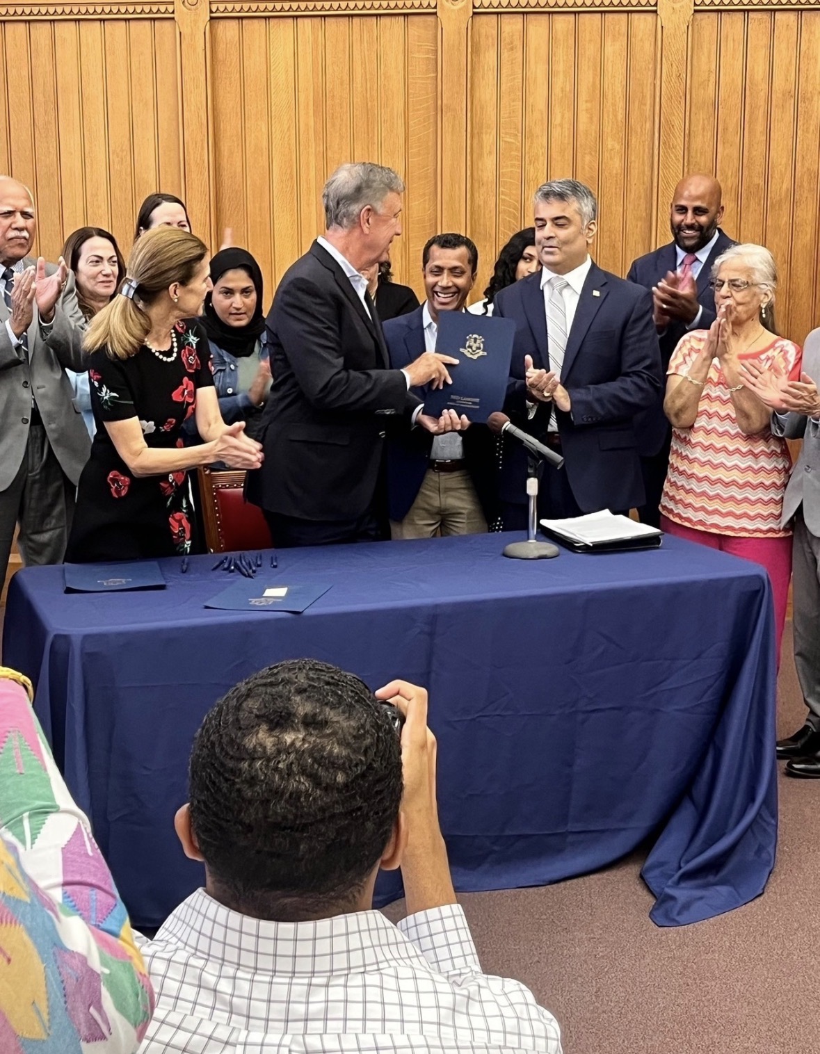 Governor Ned Lamont holding a dark blue folder beside Rajeev Pahuja as attendees applaud